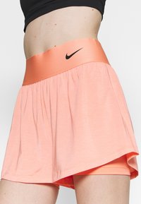 Shorts deportivos de color durazno con una amplia cinturilla naranja en contraste que presenta un pequeño logotipo negro, elaborados con un material ligero y transpirable.