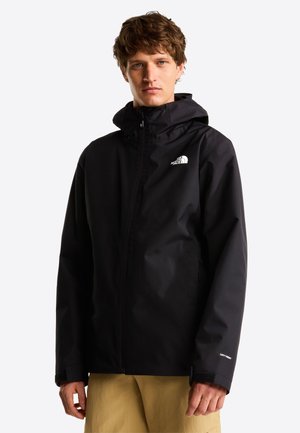 Młody mężczyzna w czarnej kurtce z kapturem North Face z suwakiem i beżowych spodniach, stojący na tle jednolitego białego tła.