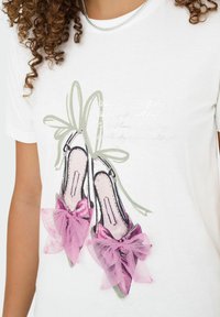 Witte t-shirt met een illustratie van roze hoge hakken versierd met grote roze stoffen strikken, gedragen door een persoon met krullend haar en een zilveren ketting.