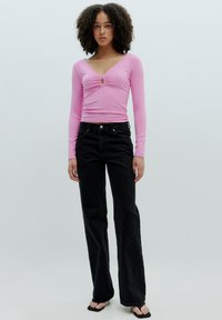 Rosa langärmliges Rippoberteil mit V-Ausschnitt und mittigem Drehdetail, kombiniert mit hochtaillierten schwarzen Schlagjeans und schwarzen Sandalen.