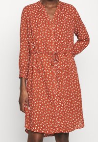 Vestido color óxido con un patrón floral, mangas largas y cinturón para atar en la cintura. Hecho de tela ligera con un ajuste suelto y detalle de botones.