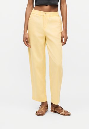 Osoba oblečená v svetložltých širokých nohaviciach s vysokým pásom, tmavom krátkom tričku a hnedých sandáloch s remienkami na plochom podpätku na bielom pozadí.
