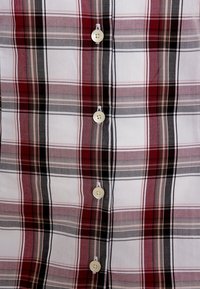 Camicia a quadri rossa e nera con righe beige e bianche, dotata di bottoni bianchi sul davanti e una texture di tessuto liscia e leggera.