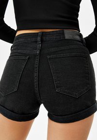 Närbild av en person som bär svarta denimshorts med rullad nederkant och en svart ribbad långärmad crop top, sedd från baksidan.