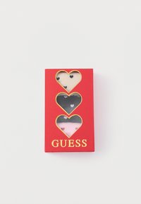 Cutie de cadou roșie cu trei ferestre în formă de inimă care arată șosete bej, negre și roz cu inimioare mici, etichetată „GUESS” cu litere aurii.