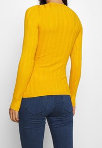 Gelber Rippenstrickpullover mit langen Ärmeln, figurbetonter Form und Rundhalsausschnitt, kombiniert mit dunkelblauen Denim-Jeans.