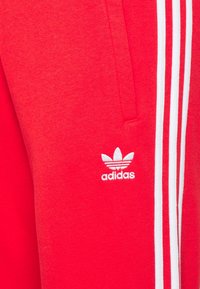 Röda Adidas-byxor med vita tre ränder och broderad Adidas-logotyp nära fickan.