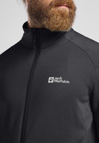 Schwarze Zip-Jacke aus strukturiertem Material, mit hohem Kragen und einem weißen Logo auf der Brust.