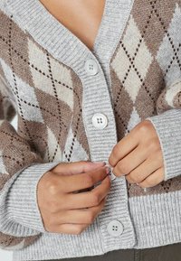 Pletený cardigan v sivej farbe s hnedým diamantovým vzorom, s rebrovanými manžetami a lemom a veľkými bielymi gombíkmi. Detail zblízka na zapínanie.