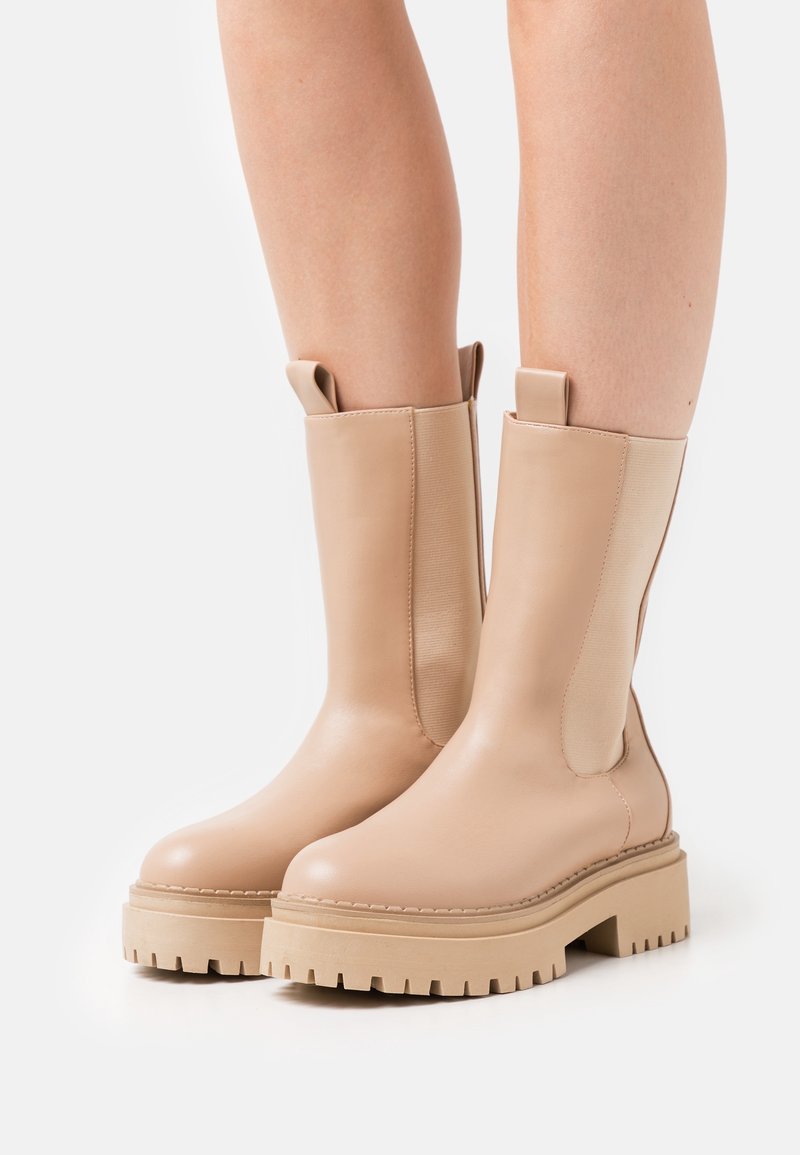 Bottes beige à semelle épaisse mi-mollet avec panneaux latéraux élastiques portées sur des jambes sur fond blanc.