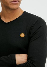 Black long-sleeve T-shirt featuring a round neckline and a small, circular orange patch on the chest with text and emblem details.