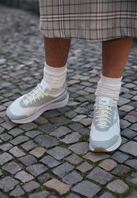 Sneakers grigie chiare con texture a rete, presentanti dettagli crema e suole beige, abbinate a calze bianche a coste su una superficie in ciottoli.