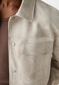 Beige mokkanahkatakki kauluksella, painonapeilla ja rintataskulla. Kangas on pehmeäpintainen ja teksturoitu.