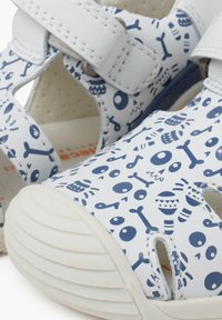 Sandalias blancas con patrones divertidos azules que presentan peces y huesos. Cierres de velcro, punta redonda y suela de goma texturizada.