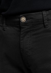 Zwarte katoenen broek met een sluiting aan de voorkant met een lichtbruine viergaatjesknop, een gladde textuur en contrasterende stiksels.