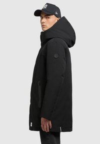 Joven vestido con un abrigo de invierno negro con capucha y una gorra de béisbol negra, de pie de lado contra un fondo claro.