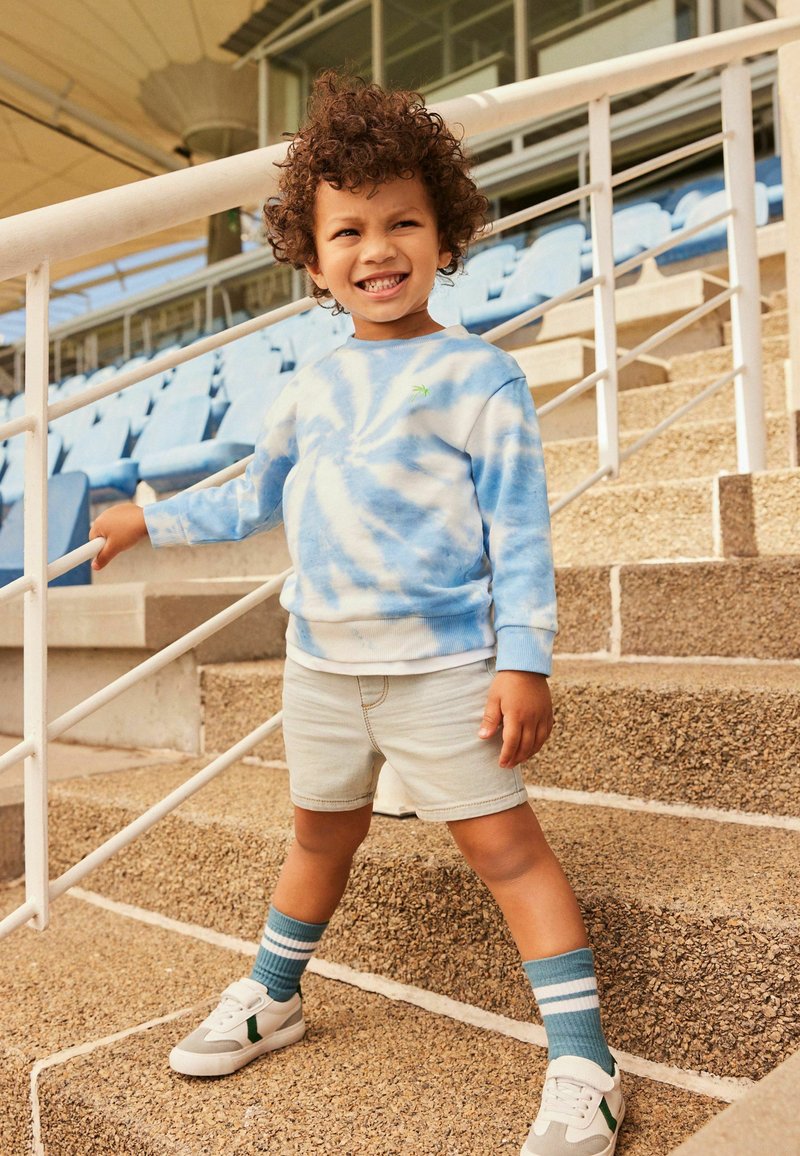 Blå tie-dye sweatshirt, ljusa denimshorts, randiga blåknee-sockor, och vita sneakers med gröna accentuerande detaljer, stående på en trappa med en hand mot räcket.