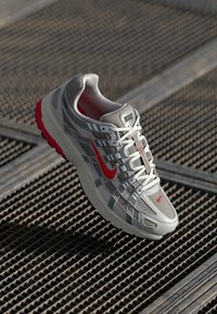 Nike Sneaker in Hellgrau mit roten Akzenten, Obermaterial aus Mesh, abgerundete Zehenpartie, dicke Sohle und strukturierte Details an den Seiten.