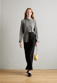 Black floor-length skirt paired with a patterned, long-sleeved shirt. Model holds a small yellow handbag with a chain strap. Black flats complete the look.