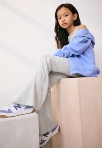 Light blue hoodie, gray flared sweatpants, and white sneakers with lavender accents. Smooth textures, simple design, seated on pastel blocks.