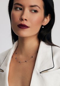 Srebrny naszyjnik z czarnymi koralikami i małym wisiorem. Modelka nosi białą marynarkę z czarnymi lamówkami i ciemnopurpurową szminkę.