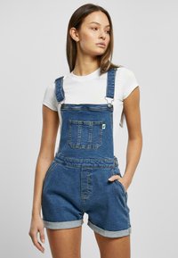 Denim overalls in a dark blue wash with a front pocket, adjustable straps, and rolled cuffs. Paired with a white shirt underneath.