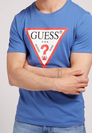 Mand med blå Guess T-shirt og arme krydset, der viser et rødt og hvidt trekantet logo med "GUESS U.S.A." og et rødt spørgsmålstegn.