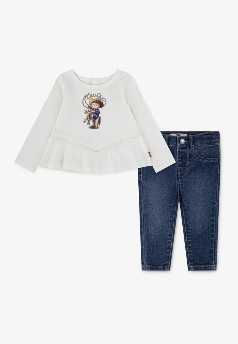 Weißes Langarmshirt mit Cowboy-Grafik und Spitzenbesatz; kombiniert mit dunkelblauer Jeans im Skinny-Fit mit vorderem Knopfverschluss.