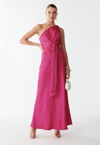 Fuksijų, vienos peties maksipalaidinės su laisvai krentančiu audiniu ir mazgo detalėmis juosmens srityje. Ji pasižymi glotnia tekstūra ir plazdančiu siluetu.