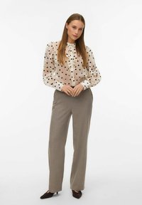 Žena stoji u prozirnoj bijeloj bluzici s crnim srcima, rukavima na naduvavanje i sivim širokim hlačama, uz crne piercing štikle.