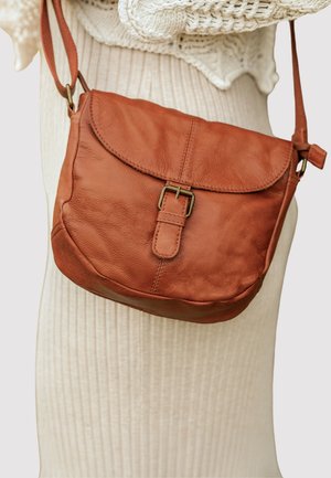 Brun læder skuldertaske med spænde på klappen, båret over en cremefarvet, ribstrikket kjole og striktrøje.