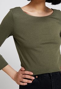 Top verde oliva a maniche lunghe con scollatura arrotondata, realizzato in un tessuto morbido e liscio. Abbinato a jeans scuri, che mettono in evidenza un design aderente.