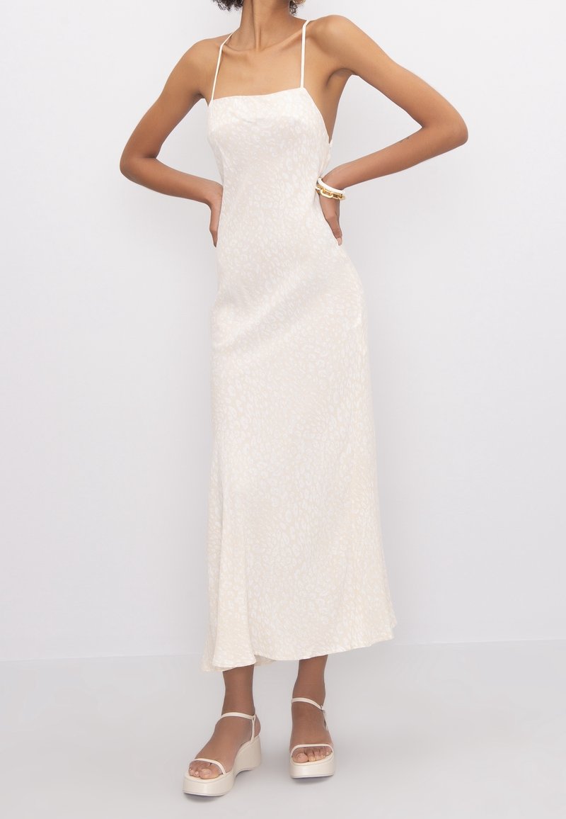 Robe de soirée blanche à imprimé léopard avec des fines bretelles, design longueur sol, texture lisse et éclat subtil. Associée à des sandales à plateforme crème.