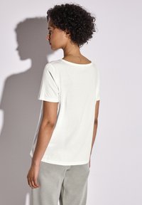Camiseta blanca de manga corta con un amplio cuello redondo, textura suave y un ajuste relajado, combinada con pantalones de lounge gris claro.