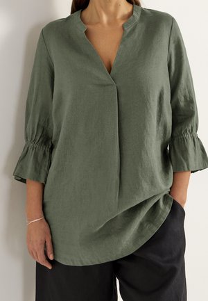 Femme portant un chemisier vert olive à col en V avec des manches cloche trois-quarts froncées et un pantalon noir à jambes larges sur un fond uni.