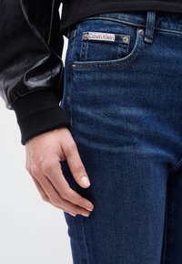 Κοντινό πλάνο ενός χεριού δίπλα σε τσέπη από μπλε τζιν με εμφανές ετικέτα Calvin Klein, φορεμένο με μαύρο μακρυμάνικο ένδυμα.