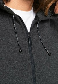 Tummanharmaa vetoketjuhuppari, jossa on teksturoitu kangas, musta vetoketju ja pyöreät huput narut. Valkoinen paita näkyy sen alla.