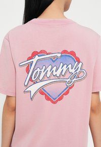 Rožinė medvilninė marškinė su širdies formos grafika, kurioje yra žodis "Tommy" mėlynu šriftu, apvadu baltos spalvos ir raudonais bangeliais.