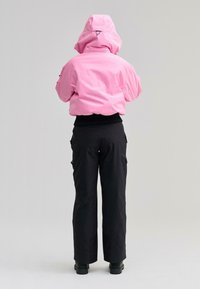 Személy élénk rózsaszín kapucnis dzsekiben, fekete nadrágban és fekete csizmában, háttal áll egy egyszerű világos háttér előtt.