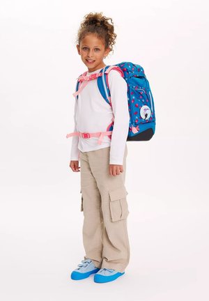 Blauer Rucksack mit pinken Punkten und Akzenten, ausgestattet mit verstellbaren Trägern und einem runden Patch. Das Model trägt ein weißes Oberteil und beigefarbene Cargo-Hosen.
