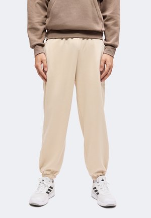 Personne portant un pantalon de jogging beige, un sweat-shirt marron et des baskets Adidas blanches avec des bandes noires, debout, les mains relâchées le long du corps.