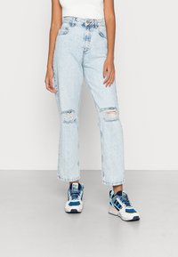 Jeans de cintura alta em azul claro, rasgados e com bordas desfiadas, combinados com tênis multicoloridos que apresentam detalhes em lona e borracha.