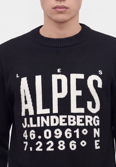 Personne portant un pull noir avec un grand texte blanc "ALPES J. LINDEBERG 46.0961°N 7.2286°E" à l'avant.