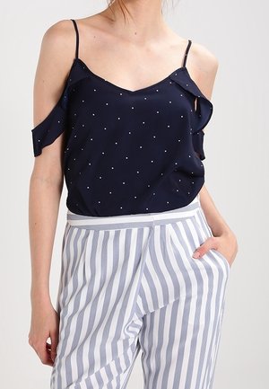 Femme portant un haut bleu marine à épaules dénudées avec des pois blancs et un pantalon blanc taille haute avec des rayures grises verticales.