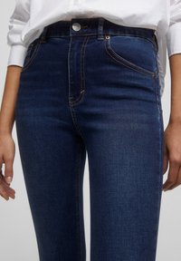 Jeans de mezclilla azul oscuro con corte ajustado, con costuras en color naranja contrastante, bolsillos frontales y cierre de botón metálico en la cintura.
