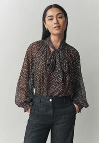 Mustmust läbipaistev pluus valgete täppidega, kaeluses lips, ja puhvis pikad varrukad, ühendatud tumedate kohandatud pükstele.