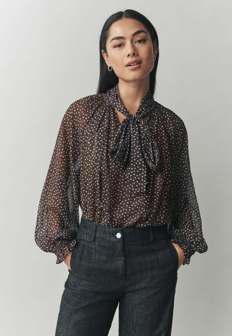 Melna caurspīdīga blūze ar baltām pērlītēm, ar loka tievu pie kakla un pūstām garām piedurknēm, saskaņota ar tumšiem pielāgotiem biksēm.