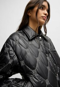 Quilted black puffer jacket featuring a large collar, snap button closure, and embroidered logos in a hexagonal pattern on glossy material.