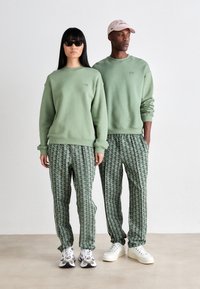 Duas modelos lado a lado, vestindo sweatshirts verdes-salvia combinando e calças de jogging verdes estampadas, uma com óculos de sol e a outra com um boné cor-de-rosa.