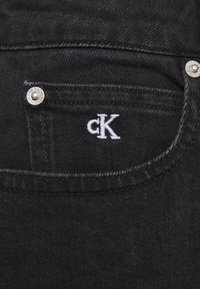 Černá denimová tkanina má přední kapsu se stříbrným knoflíkem. Nad kapsou je bílé vyšívané logo "cK".